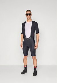 Férfi kerékpáros öltözet fekete rövid ujjú mezből hálós betéttel és sötét rövidnadrágból; fekete cipővel és napszemüveggel kiegészítve.