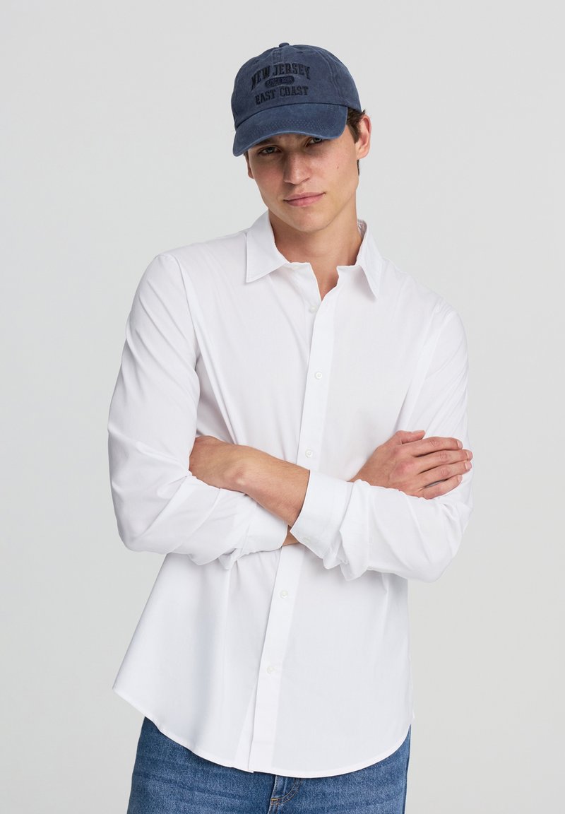 Biała koszula zapinana na guziki z klasycznym kołnierzykiem, połączona z granatową czapką baseballową z napisem "New Jersey East Coast". Casualowe dżinsy.