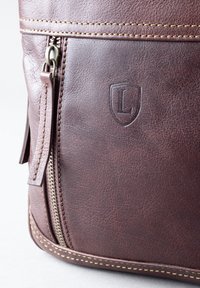 Brown leather bag with a textured finish, featuring a zippered pocket and embossed logo. Yellow stitching runs along the edges.