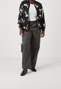 Svart bomberjacka med vitt blomstertryck, vit figursydd topp, gråvida byxor och glänsande svarta slip-on skor. Vit handväska.