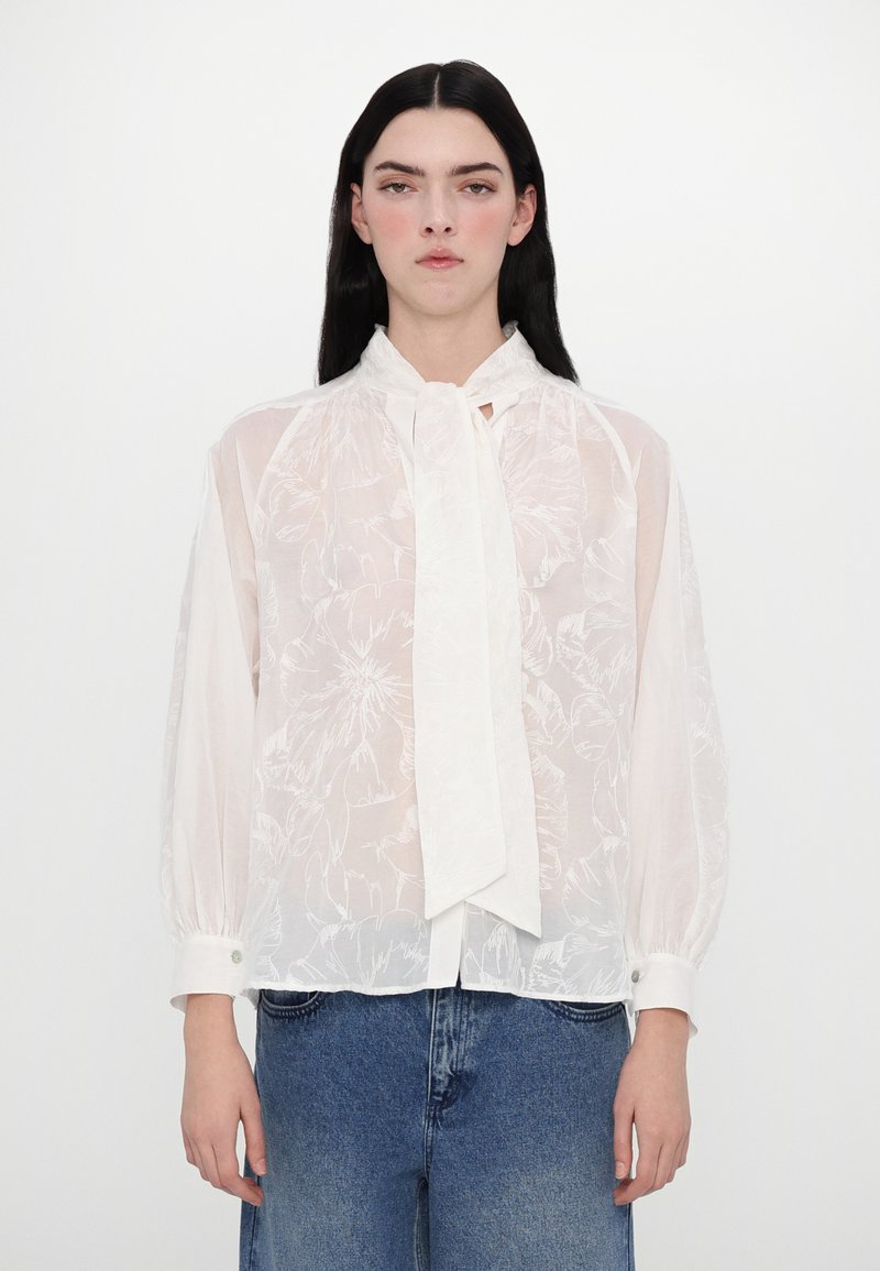 Blusa bianca trasparente con ricami floreali, collo alto e una parte anteriore annodata. Dotata di maniche lunghe con polsini bottonati e vestibilità rilassata.