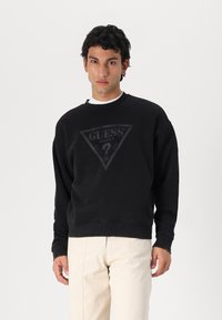 Sudadera negra hecha de tela suave, con un logo triangular que lleva el texto "GUESS" centrado en el pecho. Corte relajado y puños acanalados.