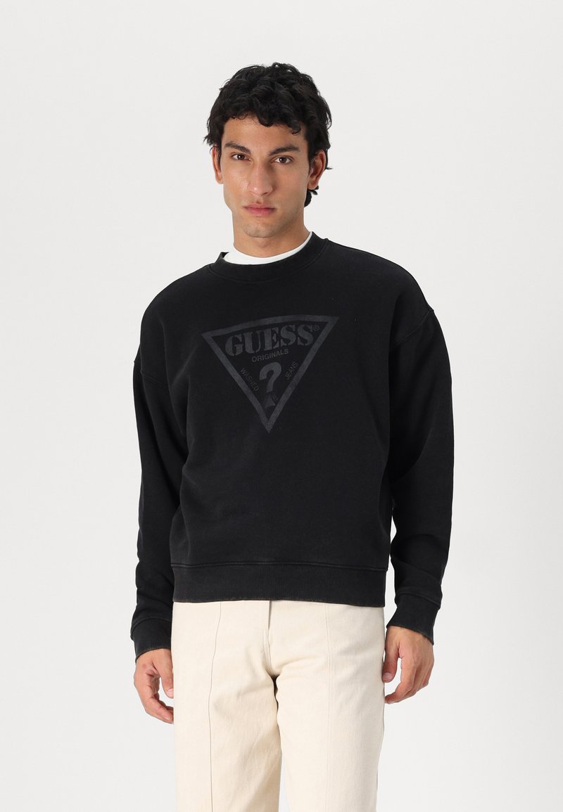 Sudadera negra hecha de tela suave, con un logo triangular que lleva el texto "GUESS" centrado en el pecho. Corte relajado y puños acanalados.