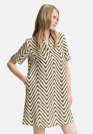 Frau mit langen blonden Haaren, die ein beiges kurzärmeliges Kleid mit auffälligen schwarzen Chevron-Streifen trägt und mit einer Hand in der Hüfte steht.