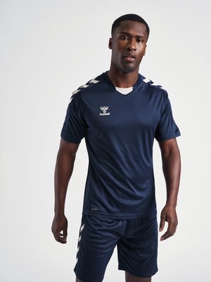 Homme debout portant une tenue de sport bleu marine avec des bandes blanches en chevron sur les épaules et le short, fond clair uni.