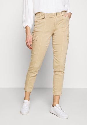 Beige cargobukser med slank pasform, der har forlommer, lynlåsdetaljer ved anklerne og blødt bomuldsmateriale, kombineret med hvide sneakers.