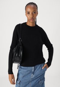 Schwarzer gerippter Pullover mit langen Ärmeln, kombiniert mit einem blauen Denim-Rock mit Seitentaschen. Eine schwarze Tasche hängt über der Schulter.