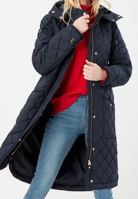Donna che indossa un lungo cappotto trapuntato blu navy, un maglione rosso e jeans blu, con una mano che tiene il colletto del cappotto e l'altra vicino alla cerniera.