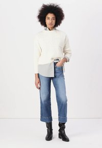 Un pull en tricot de couleur crème porté par-dessus une chemise à col bleu clair, associé à un jean bleu à jambes larges et des bottines noires.