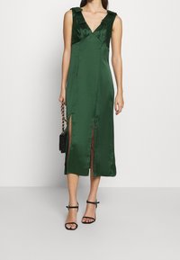 Nainen, joka on pukeutunut hihattomaan tummanvihreään satiinimidi-mekkoon, jossa on kaksi etureikää, kantaa mustaa käsilaukkua kultaketjulla ja mustia hihnakenkiä.