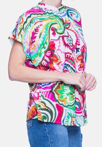 Monivärinen lyhythihainen paita, jossa on mandariinikaulus. Tässä on kukka- ja abstrakteja kuvioita vaaleanpunaisena, vihreänä ja sinisenä valkoisella taustalla.