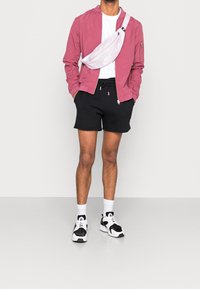 Chaqueta bomber rosa, camiseta blanca, pantalones cortos negros y zapatillas blancas con detalles negros. Presenta una riñonera rosa con un logotipo.