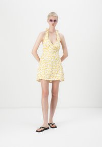 Geel bloemenjurk met een diepe V-hals en halterbandjes. Lichtgewicht stof, uitlopende silhouette en zwarte slippers. Model staat recht vooruit.