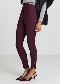 Bordeauxfarbene high-waisted Leggings mit Reißverschluss, die ein elegantes, eng anliegendes Design und seitliche Schlitze am Saum aufweisen. Kombiniert mit spitzen Absatzschuhen.