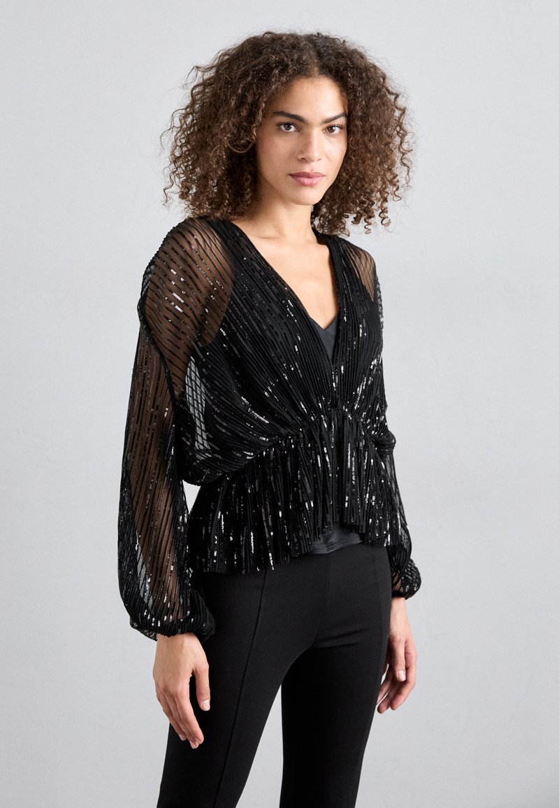 Top noir à sequins avec un décolleté en V profond, des manches en tissu transparent rayées et un design taille froncée. Porté avec des leggings noirs.