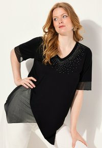 Tunique noire avec manches courtes et transparentes. Dotée d'un décolleté en V et d'ornements de petits clous brillants sur l'avant. Ourlet asymétrique.
