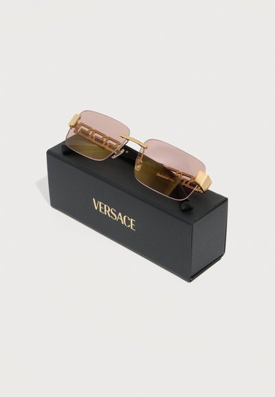 Καφέ γυαλιά ηλίου χωρίς σκελετό με καφέ διαβάθμιση και χρυσές λεπτομέρειες με μοτίβο ελληνικού κλειδιού στους βραχίονες, τοποθετημένα πάνω σε μαύρο κουτί με την επιγραφή "VERSACE" με χρυσά γράμματα.
