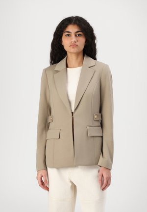 Beige blazer med en strukturert silhuett, jakkekrage, to frontlommer med klaffer og gullfargede knapper; kombinert med en hvit topp og lyse bukser.