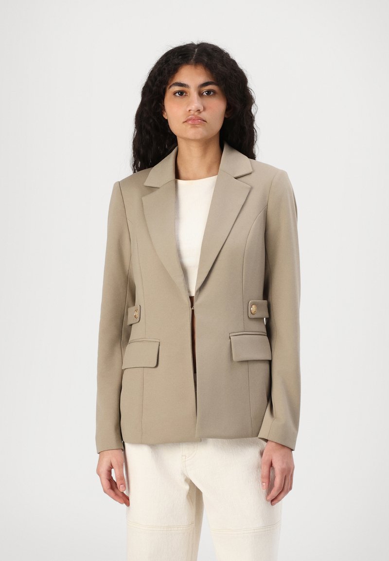 Blazer beige avec une silhouette structurée, un col à revers, deux poches avant avec rabat et des boutons dorés ; associé à un haut blanc et un pantalon clair.