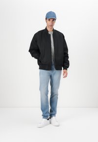 Black bomber jacket with a zip closure, gray t-shirt, light blue jeans, and white sneakers. Light blue cap completes the outfit.