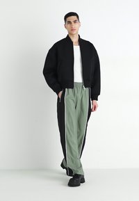 Veste bomber noire avec fermeture éclair, portée sur un t-shirt blanc. Pantalons de survêtement vert olive avec des rayures sur les côtés et une taille élastique. Chaussures noires épaisses.