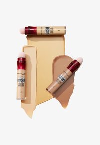 Trois correcteurs Maybelline avec des éponges arrondies, présentant des textures liquides beige clair et bronzé étalées sur un fond clair.
