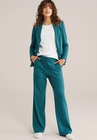 Petrolejový oblek so sako na mieru s jedným gombíkom a nohavicami s širokými nohavicami. Dopĺňa ho biela košeľa a biele tenisky. Jemná textúra.