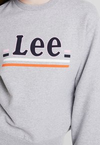 Sudadera gris con "Lee" en letras negras en negrita, acentuada con rayas rosas, blancas y naranjas debajo del texto. Textura suave, diseño casual.