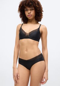 Γυναίκα με σγουρά μαλλιά που φοράει μαύρο σουτιέν με δαντελένιο τελείωμα και ταιριαστά μαύρα εσώρουχα με λεπτομέρειες από δίχτυ, στέκεται μπροστά σε απλό φόντο.