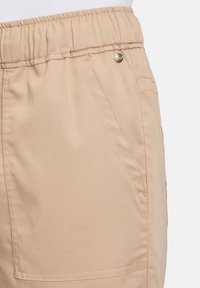 Light beige shorts with an elastic waistband, featuring a gold button detail and side pockets. Smooth fabric with a slight sheen.