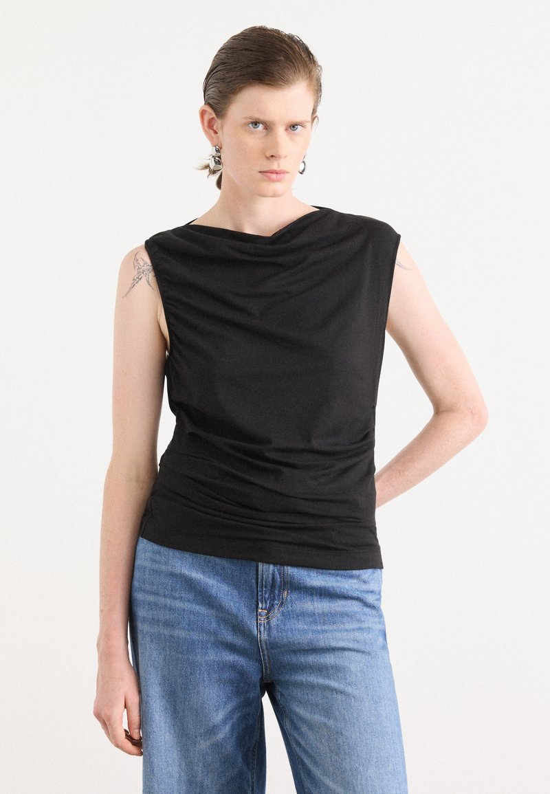 Svart ermeløs topp med drapert halslinning og rynket tekstur, kombinert med lyse denimjeans. Toppen har en glatt overflate og tettsittende design.