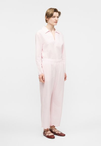Γυναίκα που στέκεται φορώντας ανοιχτό ροζ μπλούζα με μακριά μανίκια και παντελόνι φαρδύ στο ίδιο χρώμα, συνδυασμένα με μπορντό παπούτσια με κορδόνια.