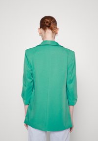 Osoba z czerwonymi włosami w kokie, nosząca złote kolczyki w kształcie kół i zieloną marynarkę z rękawami do łokcia, odwrócona tyłem w stosunku do jednolitego tła.