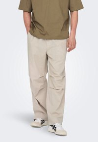 Beżowe spodnie o szerokich nogawkach wykonane z miękkiego, lekkiego materiału. Posiadają dwie boczne kieszenie i luźny krój, zestawione z białymi i czarnymi trampkami.