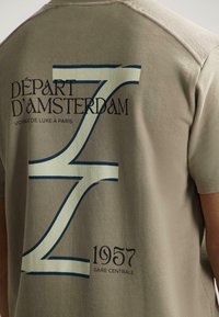 Bežinė marškinė su dideliu stilizuotu „Z“ ir tekstu „Départ d'Amsterdam“, „Voyage de luxe à Paris“ bei „1957 Gare Centrale“ nugarėlėje.