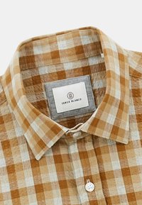 Chemise à carreaux marron et beige avec un col boutonné. Le tissu a un tissage texturé, présentant une étiquette sur un patch gris près du col.