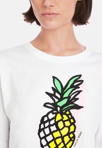 White cotton t-shirt featuring a large black outline of a pineapple, with yellow fruit and green leaves, against a light pink background.