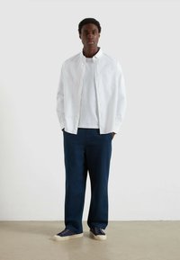 Giovane uomo in piedi con le mani in tasca, che indossa una camicia bianca a maniche lunghe aperta sopra una t-shirt bianca, pantaloni blu navy e sneakers blu navy.