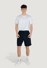 T-shirt bianca a maniche corte con logo in rilievo, abbinata a pantaloni corti blu navy con stampa del logo. Il modello indossa sneakers bianche e calze sportive.