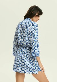 Niebieska i biała sukienka w wzory z geometrycznym motywem, z rękawami o długości trzy czwarte, w pasie z paskiem i w paskowanym kołnierzyku.