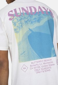 White cotton t-shirt featuring a graphic of a beach scene in blue and yellow, with "SUNDAYS" in purple text and location details below.