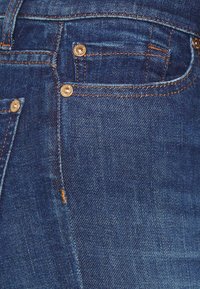 Vaqueros de mezclilla azul oscuro con costuras ámbar en contraste, remaches metálicos y un acabado texturizado; cuenta con un bolsillo delantero y pasadores para cinturón.