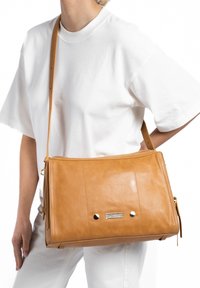 Tan leren handtas met een gladde textuur, rechthoekige vorm, verstelbare schouderriem en decoratieve hardware accenten aan de voorkant.