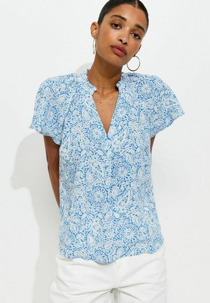 Vrouw die een blauw-witte blouse met korte mouwen en bloemmotief draagt, gecombineerd met een witte broek, en staat tegen een effen witte achtergrond.