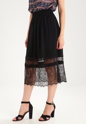 Schwarzer Midirock mit Spitzenüberzug, elastischem Bund und fließendem, transparentem Stoff. Kombiniert mit schwarzen Sandalen mit Blockabsatz.