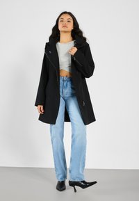 Manteau noir à capuche avec fermeture éclair à l'avant, portant un haut gris court en dessous et un jean droit bleu clair, associé à des chaussures à talons noires.