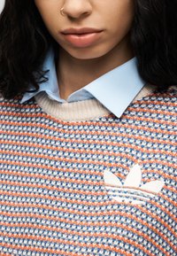 Πολύ χρωμο ριγέ πουλόβερ με κλωστές σε κρεμ, πορτοκαλί και μπλε χρώματα. Φέρει λευκό κεντημένο λογότυπο Adidas και έχει φορεθεί πάνω από ένα ανοιχτό μπλε πουκάμισο.