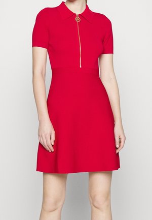 Femme portant une robe rouge à manches courtes avec un col et une fermeture éclair avant ornée d'un pendentif circulaire en or avec le logo MK.