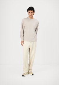 Beidzīga krekls ar garām piedurknēm, krēmkrāsas bikses ar brīvu piegriezumu un bēšas kedas. Minimālistisks dizains ar smalku zīmolu.