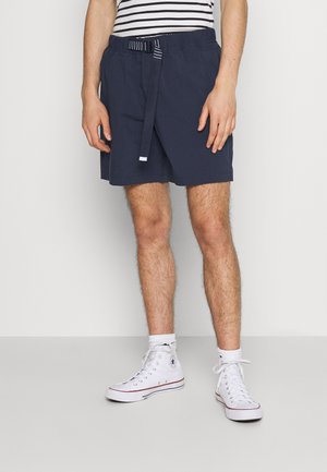 Pantaloncini navy con vita elasticizzata e cintura a righe, realizzati in tessuto leggero. Indossati con sneakers bianche alte e calzini corti.